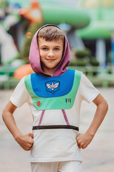 A young boy is wearing a purple hood attached to a white t-shirt. There is blue and green on the chest of the shirt with purple lines and a black band along the waist resembling an astronaut. There is an embroidered white symbol, red circle and red, blue and green lines that resemble space suit buttons.