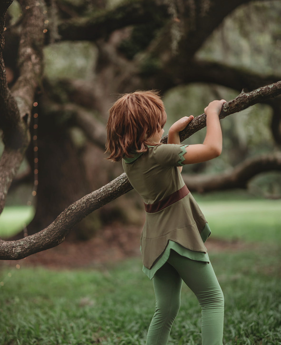A young child is wearing an earthy green tunic with leaf green collar. The short sleeves have a double layer detail. The bottom hem is a handkerchief cut.