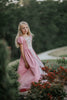 A young girl wearing a pink Sleeping Beauty Princess Maxi Dress. Short puff sleeves. A lighter pink panel is on the chest with gold rick rack criss crossed accent over it. The long dress is a soft cotton.