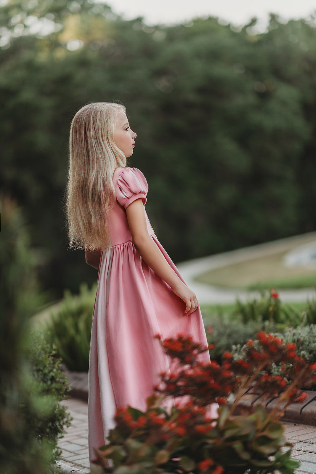 A young girl wearing a pink Sleeping Beauty Princess Maxi Dress. Short puff sleeves. A lighter pink panel is on the chest with gold rick rack criss crossed accent over it. The long dress is a soft cotton.