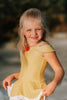 A young girl with long blonde hair is standing on the beach, wearing a yellow swimsuit with white trim and a red rose detail from the Red Rose Princess Collection. The 2 piece swimsuit is reminiscent of a princess ball gown and is SPF 50 for sun protection.