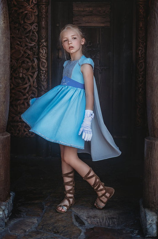 A young girl in an Ice Queen Dress and Cape Set, with a sheer chiffon removable cape. The light blue cap sleeves have white lace over them and silver racking on the bottom. There is a silver sparkle panel on the chest and a silver embroidered snowflake on the darker blue fabric on the chest. Silver trim lines the bottom of the light blue twirl dress.