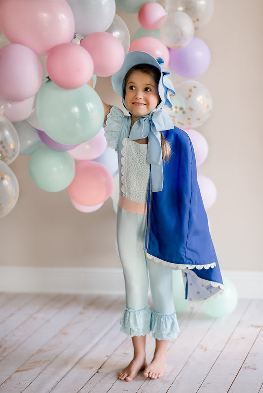 A young child dressed in the Bo Peep Romper and royal blue Cape (removable). A light blue romper adorned with white lace trim, and leggings with ruffled ends. The flutter sleeves are eyelet lace.