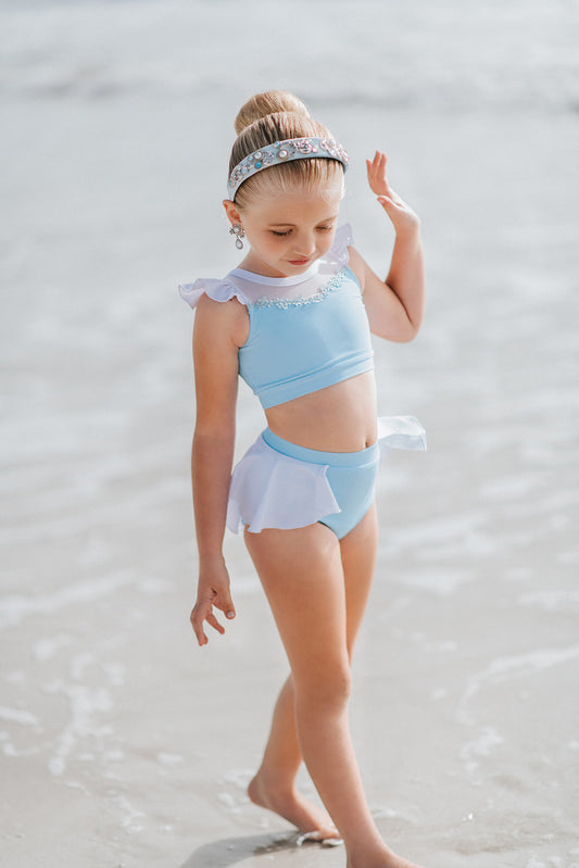 A young girl is wearing a 2 piece light blue and white swim suit. The top has ruffles on the shoulder and light blue floral embroider at the neckline. The modest bottoms are light blue with a white skirt that wraps around the back and is open in the front.