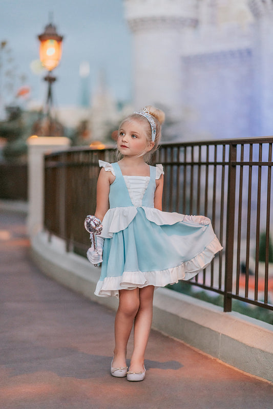 A young girl in an "Our Original Cinderella Twirl Dress" stands near a castle. The dress has a white flutter ruffle on the straps. There is a white skirt ruffle that overlays the blue twirl skirt. The soft cotton material is comfortable and breathable.