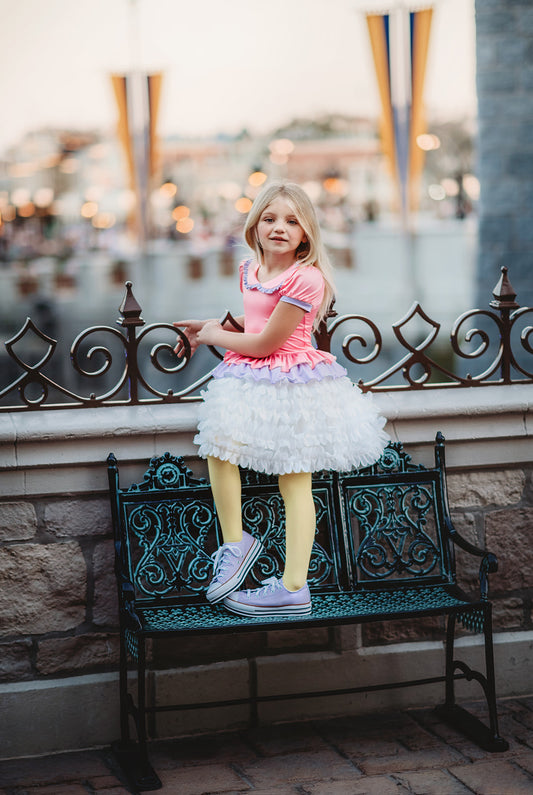 A young girl is wearing true pink and lavender bodice and white petal skirt dress. The top has puff sleeves and ruffled collar. The white skirt imitates feathers and is lined with tule and cotton to give a fuller appearance.