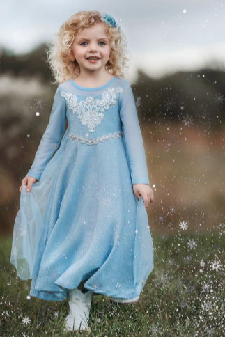 A young girl is dressed in the sparkly light blue Portrait Collection Ice Queen Gown adorned with intricate custom lace at the neckline with rhinestones. A light blue cape with silver sparkle snowflakes is buttoned to the back of the gown.