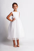A young girl in a Pearl Accent gown in white, featuring a bow at the back. Woven cotton bodice and large pearl beads line the neck and back edge! A full tulle skirt with tea length hemline and modest open back.