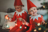 A young child dressed in Unisex Elf Pajamas with a red pointy hat. The perfect Christmas photo outfit. The fabric is bright red and buttery soft. The top is long sleeved with white cuffs and a white collar that has large triangle points similar to a clown. Long pants with white cuffs.