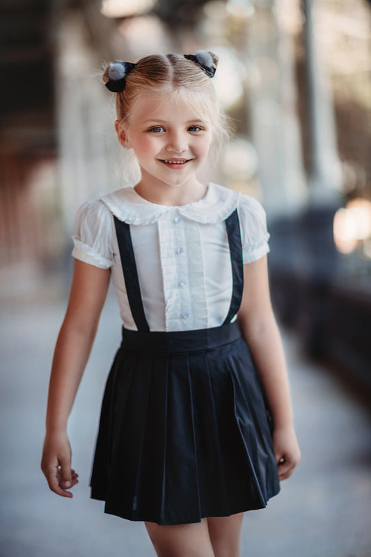 A young girl wears the Plaza Girl Everyday Dress—a white button-down shirt with ruffle accents and short sleeves tucked into a knee-length black skirt with suspenders made of high-quality woven cotton.