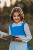 A young girl stands wearing the Red Rose Princess Village Dress from the Fairy Godmother Collection, reminiscent of Disney's Belle from "Beauty and the Beast." The white sleeves are long. The dress has a bight blue chest and skirt. There is a white apron at the waist and white embroidery lace at the base. Dress is mid calf.
her 100% woven cotton dress to flare out. The background is softly blurred with trees and sky visible.