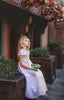 A young girl is wearing the light purple Rapunzel Maxi Dress. The lavender princess dress features puffed sleeves and a crisscrossed gold trim. The light pink panel adorns the middle front and back.