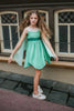 A young girl in a leaf green twirl dress. Sleeveless with nude mesh neckline. Detachable fairy wings flow openly or can be held by finger holes. Handkerchief style hemline for the true fairy feel.
