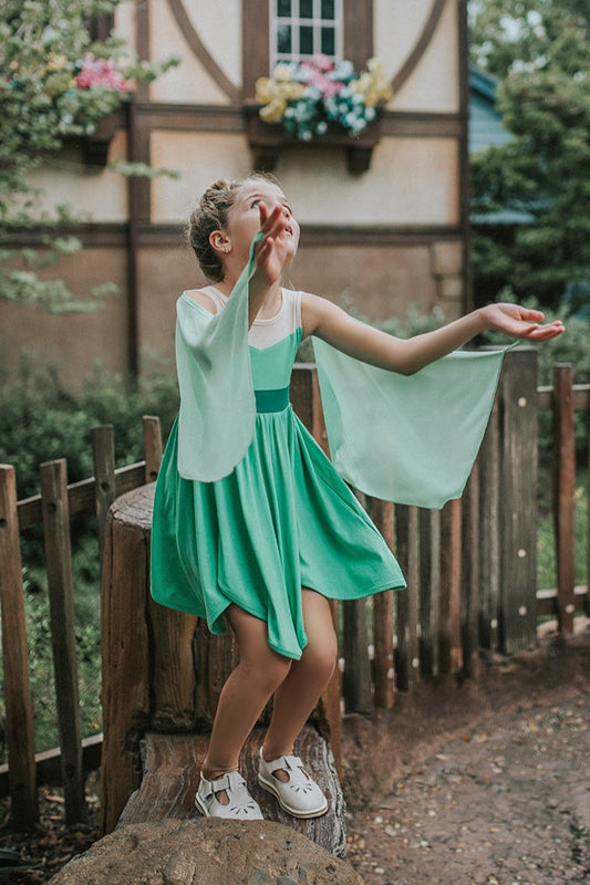 A young girl in a leaf green twirl dress. Sleeveless with nude mesh neckline. Detachable fairy wings flow openly or can be held by finger holes. Handkerchief style hemline for the true fairy feel.
