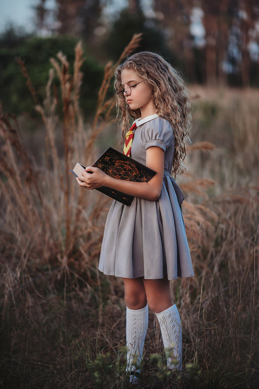 A young girl is wearing a grey Wizard Girl Twirl Dress with a striped yellow and brick red tie. The twirl dress has a white collar, short puff sleeves and a tie in the back at the waist.