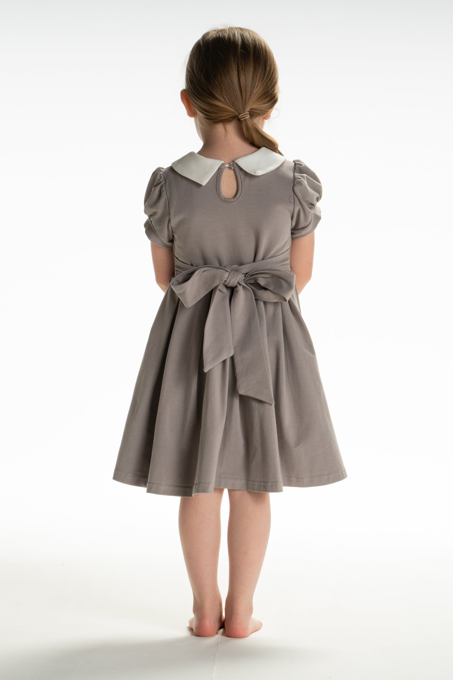 A young girl is wearing a grey Wizard Girl Twirl Dress with a striped yellow and brick red tie. The twirl dress has a white collar, short puff sleeves and a tie in the back at the waist.