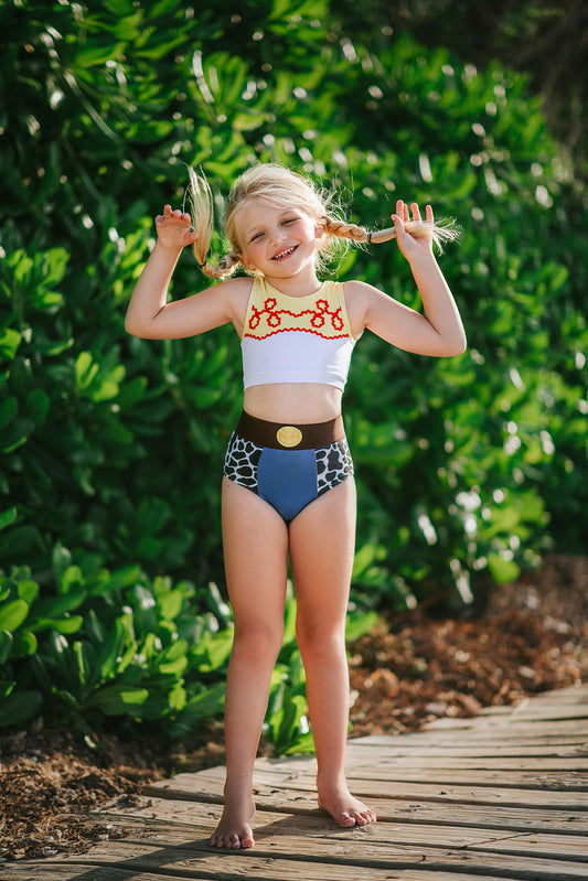 A young girl is wearing a 2 piece swim set. The top is white with yellow on the upper chest. Across the yellow is red scrolling rick rack. The bottoms are a faux denim with black and white cow print panels.