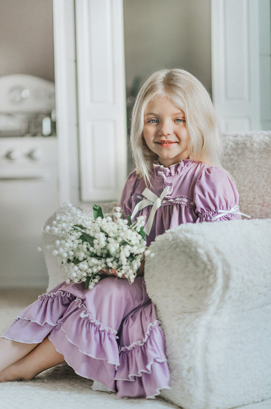 A young girl is wearing The Original Clara Nutcracker Gown in Lavender, featuring lavender ruffles and a white bow. The buttery soft fabric is cozy and sensory friendly. The puff sleeves with ribbon detail and the ruffles at the hem add a traditional classic look.