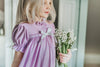 A young girl is wearing The Original Clara Nutcracker Gown in Lavender, featuring lavender ruffles and a white bow. The buttery soft fabric is cozy and sensory friendly. The puff sleeves with ribbon detail and the ruffles at the hem add a traditional classic look.