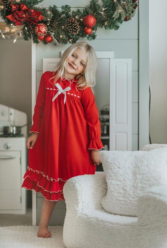 A young girl is wearing The Original Clara Nutcracker Christmas Gown Long Sleeve in Red, adorned with a white bow. There are two buttons on the next with a ruffle at the top. There is one ruffle at the bottom of the sleeve and two ruffles at the bottom of the gown. Buttery soft polyester knit fabric.