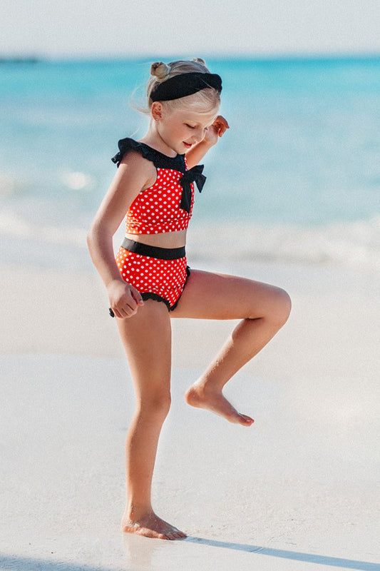 A young girl is wearing our Miss Mouse 2pc Swim set. This 2pc swim has SPF 50. The top is red with white polka dots. There is a large black bow in the center and small ruffles on the shoulder. The bottoms have black ruffles along the edge.