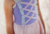 A young girl stands on a beach wearing a pastel pink and purple Rapunzel One Piece Swimsuit, adorned with crisscross and lace detail. The bodice is lavender and skirt is light pink. The swimsuit is SPF 50 for protection from the sun.