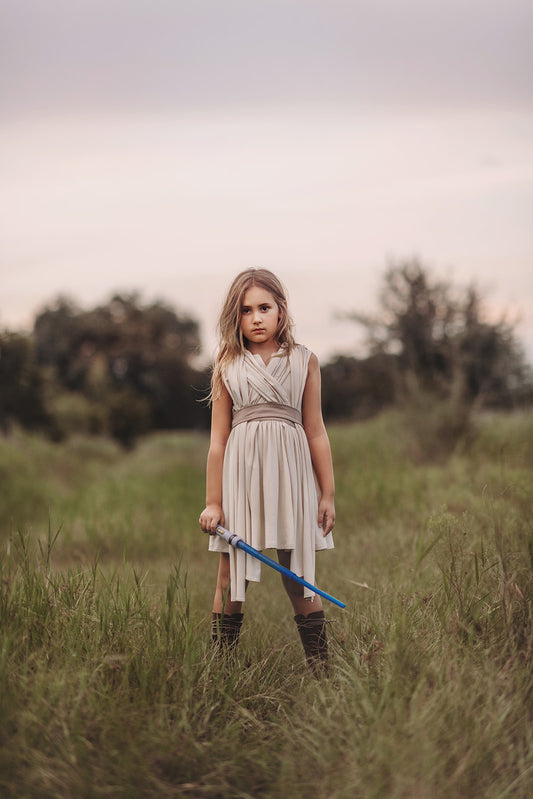 In a far away galaxy a girl and her lightsaber stand in a beige dress. There are layers of fabric that cross and intersect on the bodice. The cotton twirl dress ties at the waist in the back.