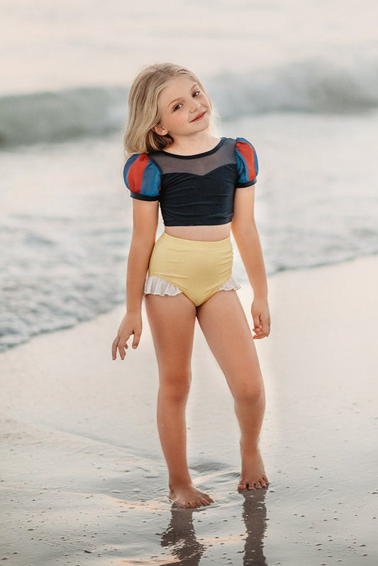 A young girl is wearing the Snow White 2 piece Swim Set with SPF 50 for sun protection. The navy blue top has blue and red puff sleeves and navy mesh at the top. The light yellow bottoms have a white ruffle on the leg.