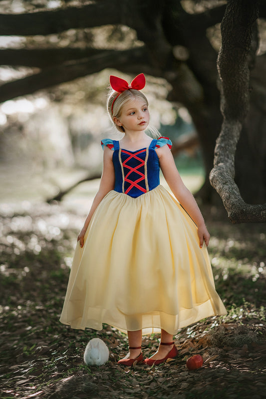 A young girl stands in a forest wearing the Portrait Collection Snow White Gown. The gown is velvet blue and a light yellow skirt with a red ribbon criss cross accent on the bodice. The dress is sleeveless with a red and blue flutter ruffle on the top.