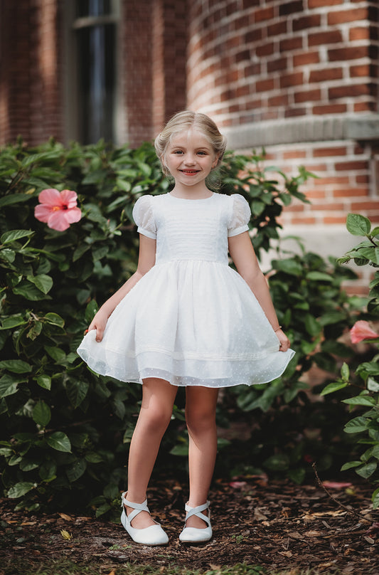 A young girl is wearing our Swiss Dot Party Dress. The elegant white dress has puff sleeves and a white lace trim on the hem.