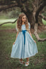 A young girl in the Lost Child Blue Gown with puffed sleeves and a blue ribbon tied at the waist. There is lace at the bottom of the sleeves and in two vertical stripes on the chest.