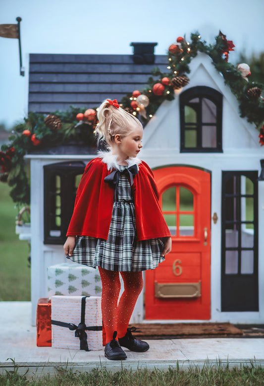 A young girl stands dressed in a black and white vintage plaid dress. A red velvet cape trimmed with white fur and a black bow are tied around her shoulders. The dress is mid thigh length and a sewn in pettiskirt add fullness to the bottom of the dress.