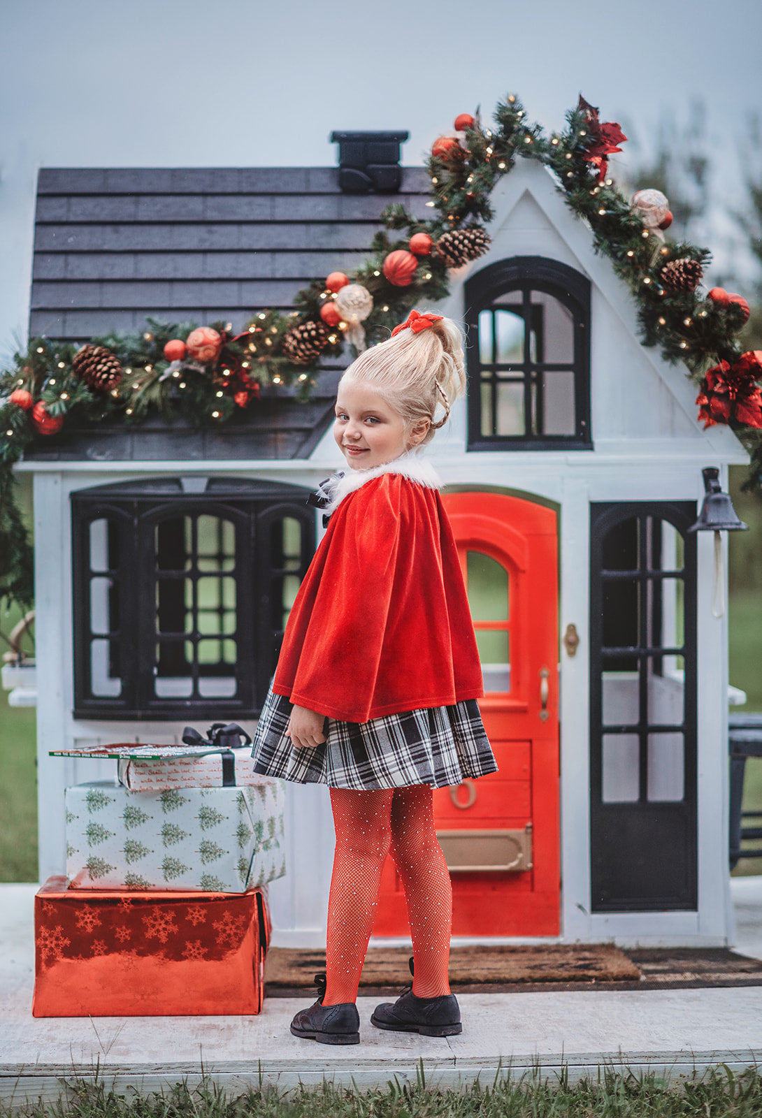 A young girl stands dressed in a black and white vintage plaid dress. A red velvet cape trimmed with white fur and a black bow are tied around her shoulders. The dress is mid thigh length and a sewn in pettiskirt add fullness to the bottom of the dress.