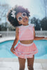 A young girl is wearing a 2 piece pink swim set. There is white collar with a pink bow at the neck. Two lighter pink stripes are on the chest like ribbon tails. The bottom is a skirt with bows light pink bows at the hem.
