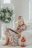 A young child with curly blonde hair stands barefoot on a soft carpet, holding an open book. The child wears *The Original Clara Nutcracker Gown in Winterberry* and reads intently. A decorated Christmas tree is visible in the background, part of our exclusive Christmas Lounge Collection.