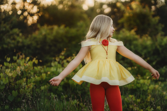 A young girl stands outdoors in a grassy area with trees in the background. She is wearing a Red Rose Princess Peplum Top from the Only Little Once Exclusive Collection, adorned with a red flower embellishment, and red leggings. The light behind her gives the surroundings a warm, glowing effect.