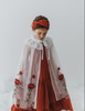 A young girl is wearing a red dress with a white tulle cape adorned with embroidered red roses. The floral details of her ensemble are striking.  The shades of red and green are the perfect addition  to our Red Rose Princess Collection.