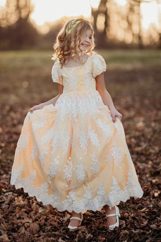 A young girl is dressed in the Red Rose Regency Gown, a pastel yellow with white lace overlay. The puff sleeves add a the princess effect. The empire waist dress adorned with white floral patterns and complemented by a matching Red Rose floral headband. 