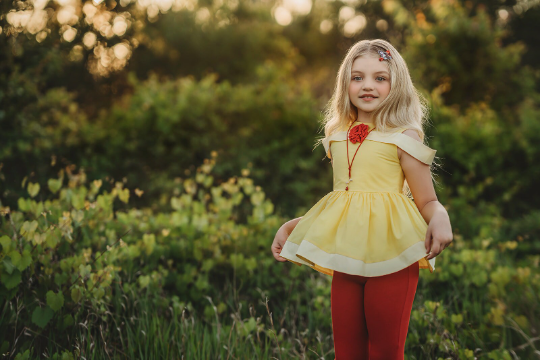 A young girl stands outdoors in a grassy area with trees in the background. She is wearing a Red Rose Princess Peplum Top from the Only Little Once Exclusive Collection, adorned with a red flower embellishment, and red leggings. The light behind her gives the surroundings a warm, glowing effect.