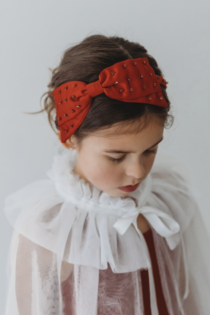 A young girl is wearing a red dress with a white tulle cape adorned with embroidered red roses. The floral details of her ensemble are striking.  The shades of red and green are the perfect addition  to our Red Rose Princess Collection.