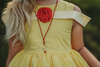 A young girl stands outdoors in a grassy area with trees in the background. She is wearing a Red Rose Princess Peplum Top from the Only Little Once Exclusive Collection, adorned with a red flower embellishment, and red leggings. The light behind her gives the surroundings a warm, glowing effect.