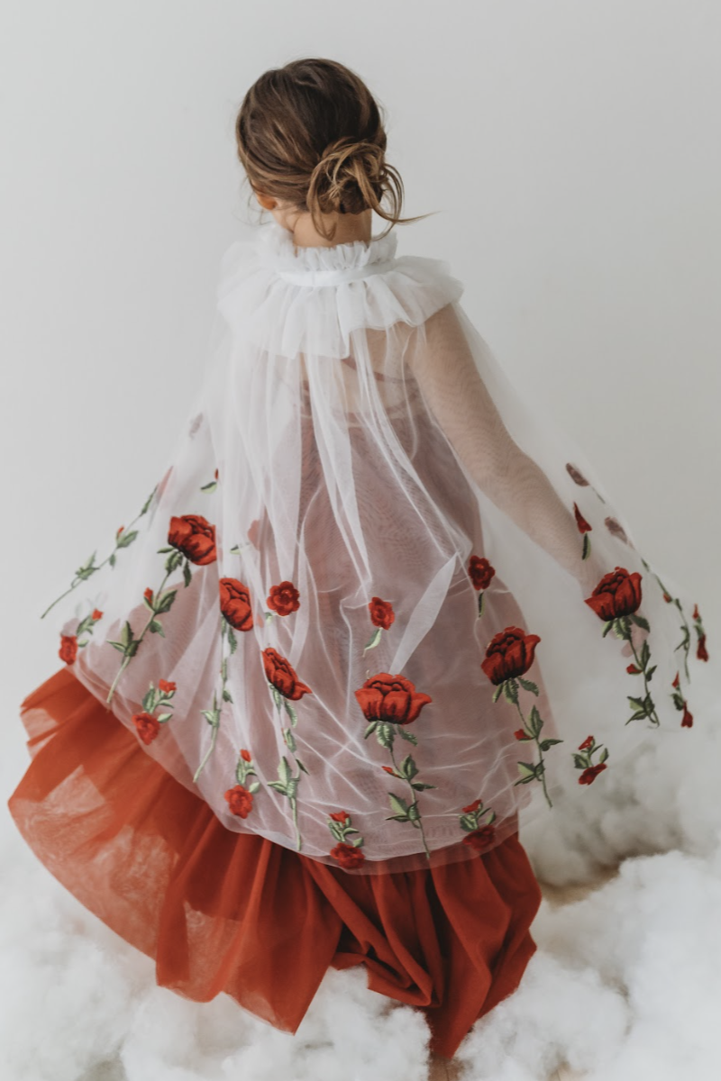 A young girl is wearing a red dress with a white tulle cape adorned with embroidered red roses. The floral details of her ensemble are striking.  The shades of red and green are the perfect addition  to our Red Rose Princess Collection.