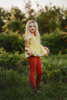 A young girl stands outdoors in a grassy area with trees in the background. She is wearing a Red Rose Princess Peplum Top from the Only Little Once Exclusive Collection, adorned with a red flower embellishment, and red leggings. The light behind her gives the surroundings a warm, glowing effect.