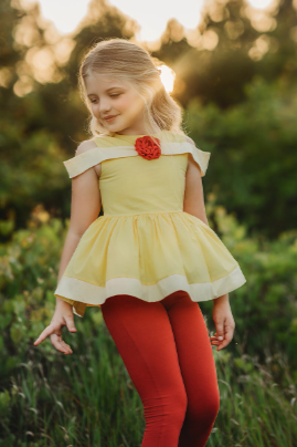 A young girl is wearing a Red Rose Princess Peplum Top adorned with a red flower embellishment. The off the shoulder cut is reminiscent of a ball gown.  The peplum bottom has tulle for fullness.  The cotton is soft and comfortable. 