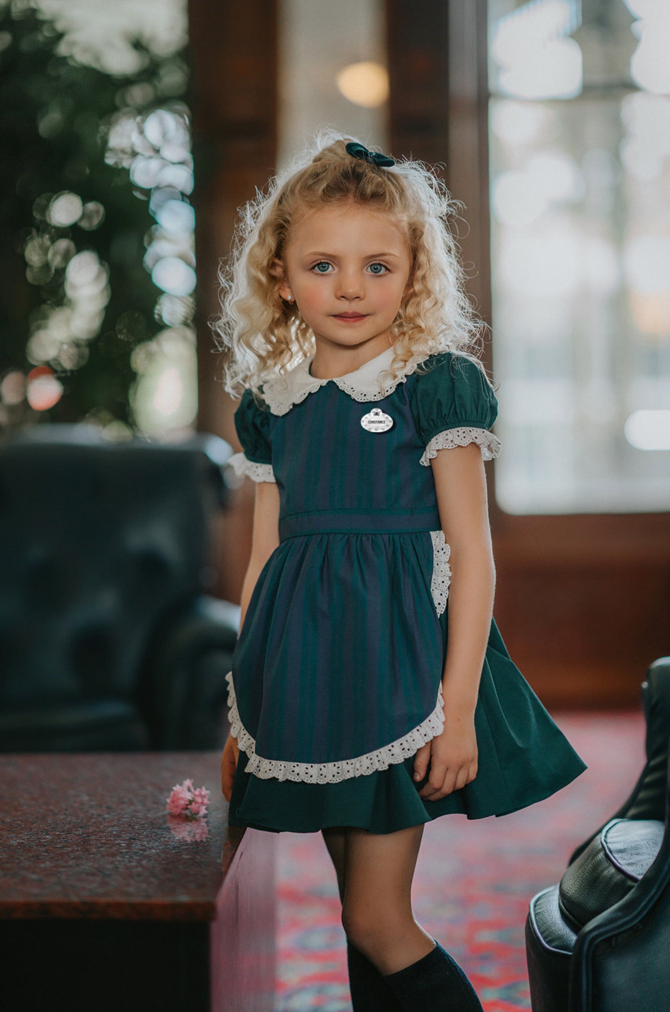 A young girl stands indoors wearing a dark teal green dress with white lace trim, a matching plaid blue and green apron, puff sleeves and a white collar.  The woven cotton is perfect for spooky season.