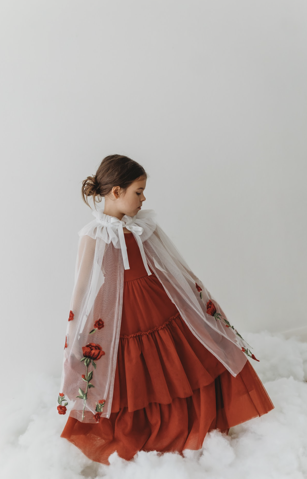 A young girl is wearing a red dress with a white tulle cape adorned with embroidered red roses. The floral details of her ensemble are striking.  The shades of red and green are the perfect addition  to our Red Rose Princess Collection.