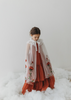 A young girl is wearing a red dress with a white tulle cape adorned with embroidered red roses. The floral details of her ensemble are striking.  The shades of red and green are the perfect addition  to our Red Rose Princess Collection.