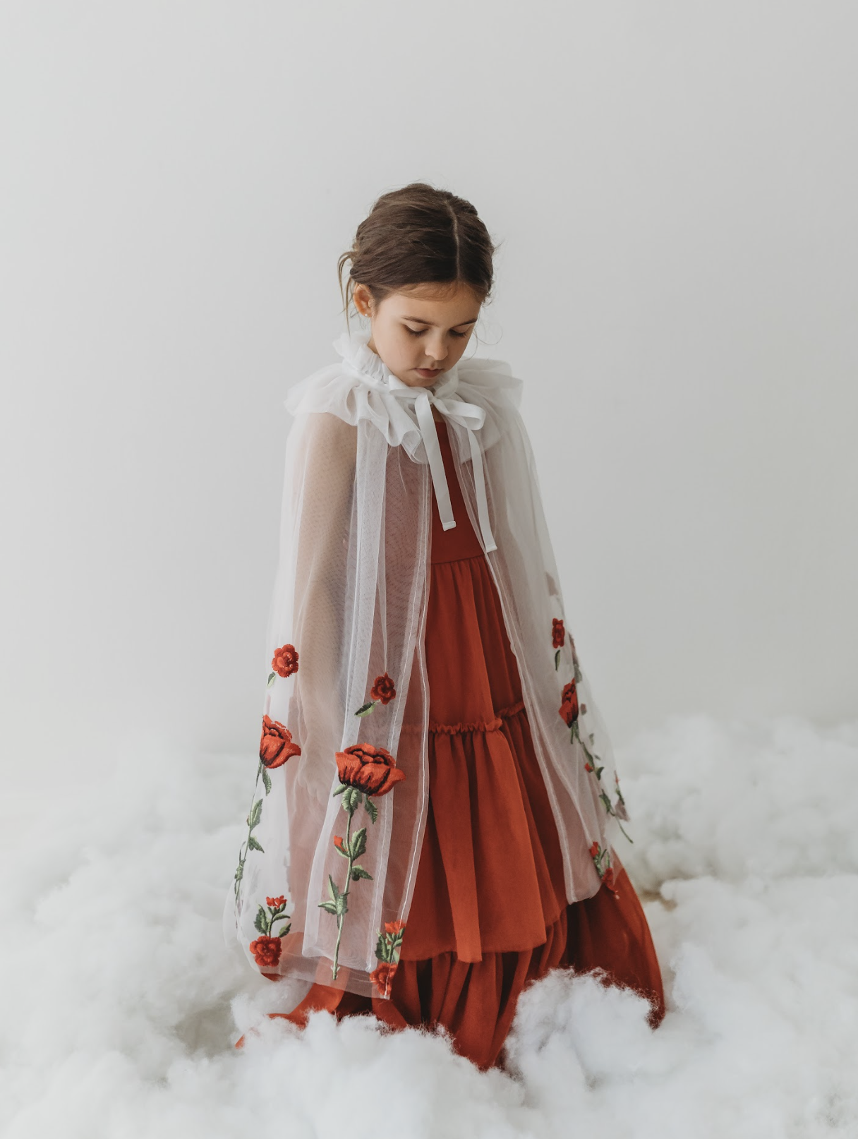 A young girl is wearing a red dress with a white tulle cape adorned with embroidered red roses. The floral details of her ensemble are striking.  The shades of red and green are the perfect addition  to our Red Rose Princess Collection.