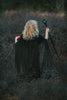 A person with curly blonde hair, seen from behind, wears the Malevolent Mistress Maxi dress and Cape Set that glimmers like glitter tulle and holds a glowing green staff in one hand and a red orb in the other. They stand in an overgrown, twilight forest, evoking a magical and mysterious villain costume atmosphere.