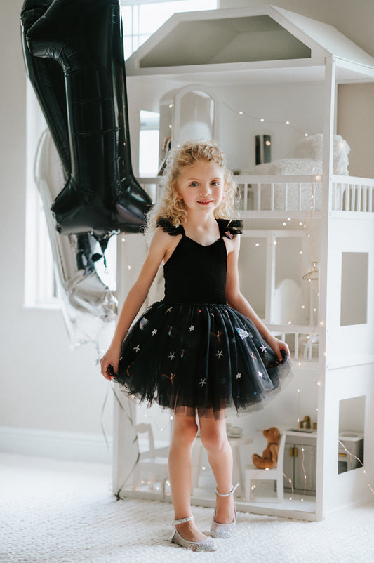 A young girl is wearing our Space Party Tutu.  The black tutu dress has a velvet bodice with tule ruffles on the straps.  The skirt is black tulle with sequin shapes of stars, planets, comets and more. 